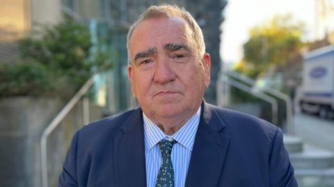 A mid shot of Simon Vermeulen. He has short brown hair and is wearing a navy jacket, strip shirt and tie.