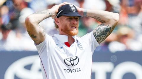 Ben Stokes, wearing a blue England cap, stands with his hands on his head