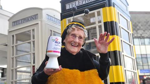 Jean Bishop wearing glasses and dressed in her bee outfit stands in front of a telephone box painted black and yellow. She is holding a white Age UK collecting tin.