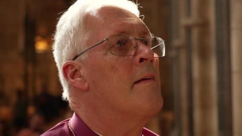The Bishop of Southwark, the Right Reverend Christopher Chessun in clerical maroon robes in Southwark Cathedral. He has white hair and is wearing glasses looking upwards - there are no obvious signs of scarring.