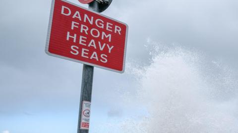 A red-coloured roadsign reading 'Danger from heavy seas', against the backdrop of a cloudy sky. Seawater spray can be seen alongside the sign.