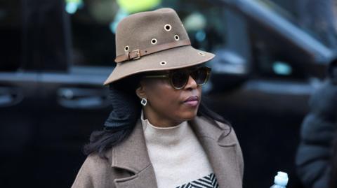 A woman wearing a hat, glasses, a coat and sweater is seen from the side