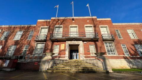 A red brick building with white window frames and three flag poles on the roof. 