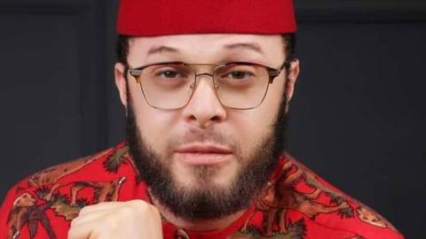 A head shot Alex Ikwechegh - with a beard and glasses - in a red hat and red shirt with animals on it