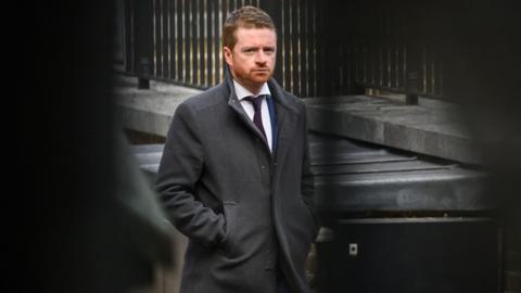Morgan McSweeney, the prime minister's former chief of staff, walking down Downing Street. He has a serious expression and wears a dark grey coat with the collar turned up over a white shirt with a black tie. He has short hair and a very short beard.
