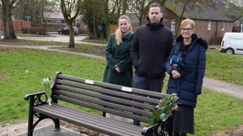Emma Callaghan has long blonde hair in a ponytail and is wearing a long green wool coat. She is standing next to Gavin Callaghan, who is wearing a black jacket and grey trousers. Patricia Reid is wearing a navy puffer coat with a fur hood and a black and silver outfit underneath. She has an arm in a blue sling and it is strapped around her neck. They are standing on a grassed area behind a bench with a bunch of flowers on each side.