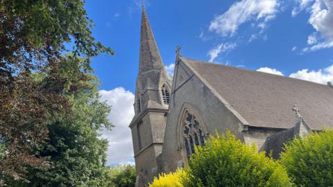A view looking upwards at a stone church and its church spire. There are lime green bushes in the foreground and above is a blue sky with white clouds.