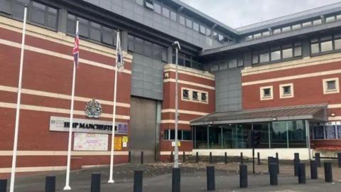 A view of the front of HMP Manchester, which is still widely known by its former name of Strangeways. On the right of the photograph is the jail's glass-panelled entrance. In the middle of the picture there is a vehicles' entry point - it has silver-coloured metallic doors which are two storeys in height.