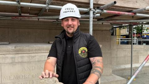 The photo shows a man smiling at the camera. He's wearing a black gilet, back polo shirt and white hard hat. He's standing in front of a warm memorial, and has his hand on a piece of stone. The memorial is covered in scaffolding.