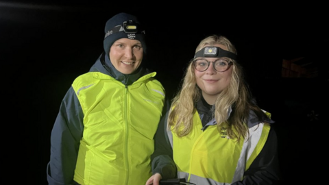 A woman with a black hat and black coat alongside a woman with long blonde hair and glasses. Both are wearing yellow reflective jackets