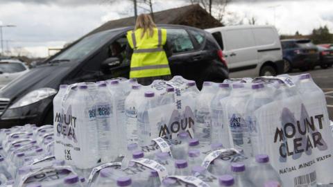 Packs of bottled water stacked up in focus while a woman in a yellow high vis vest talks to someone in a car. 