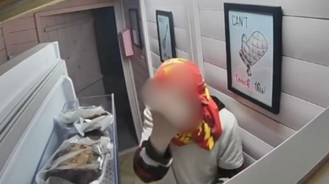 An image from CCTV shows what appears to be a young man with a red and yellow football shirt wrapped around his head like a balaclava. He has opened up a fridge with cakes in wrappings in the foreground. The person's hand has been blurred out, as he makes a rude gesture at the cctv.