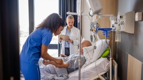 A hospital patient lying in a bed is being tended to by a nurse wearing a blue uniform, and a doctor in a white coat is standing behind the bed, in front of a window
