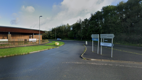 A picture of the road where the collision took place. It is a large road just off of a roundabout. A wooden building can be seen to the left of the screen with road signs in the middle.