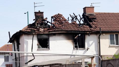 The roof of the property is badly burnt. There are pieces of wood or metal sticking up out of it. The holes where the windows were are blackened.