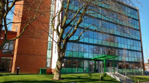 The front of North Hertfordshire Council's building in Letchworth Garden City. There are two trees without leaves in front of the building, the building itself is six stories high and has a lot of windows with a set of steps leading towards its entrance. The sky is blue behind the building.