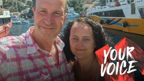 Neal and his wife Tara are standing in front of some water, with ferries behind them and a hilly village on the other side of the bay.