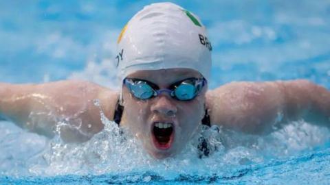 Swimmer Dearbhaile Brady mid-stroke, facing the camera, taking a breath. She is wearing reflective goggles and a swimming cap.