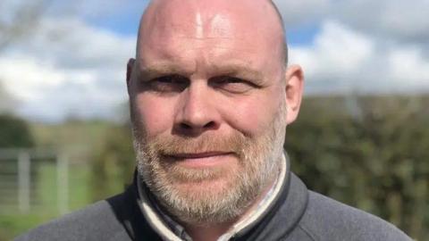 A bald man with a beard, wearing a grey jacket.