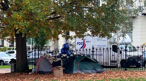 Three tents, surrounded by leaves, a bunch of blue and silver balloons, and a washing basket, sit beneath a tree at the edge of a Cheltenham park, bordered by wrought iron fencing. White Regency style terraced homes can be seen on the other side of the road, where cars and a van are parked.