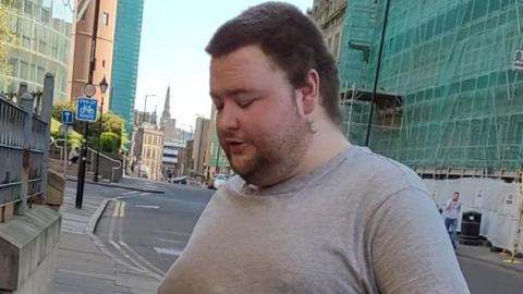 A man with short brown hair and a beard wearing a grey top pictured in a street