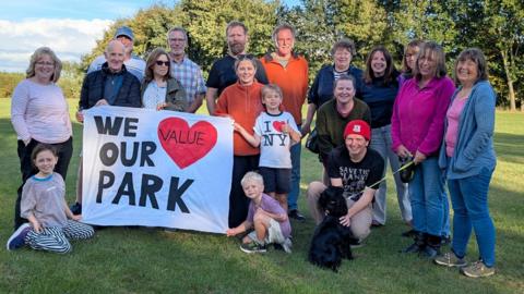 A mixed age group of residents - children and adults - from North Cowton village stand in a open green space. Several people hold up a white sign with black lettering and a red love heart which reads: 'We Value Our Park'.