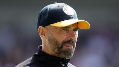Paul Warne in a baseball cap looks on during a MK Dons match