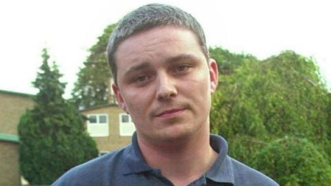 Ian Huntley has cropped brown hair. He has little expression on his face and has tilted his head slightly. He is wearing a navy polo shirt. Several trees are behind him in this picture taken in 2002.