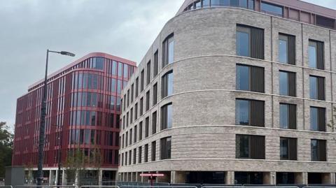 A modern curved, grey-brick building stands in front of a modern red clad, curved building. A lamppost and some small trees can be seen. The sky above is full of thick, grey cloud.