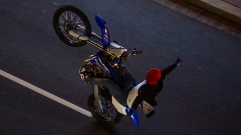 Aerial shot of a dirt bike with the rider doing a wheelie and holding out one arm. He wears a red balaclava