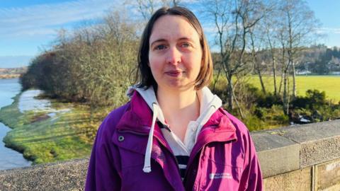 Charlotte Davys standing on a bridge and looking at the camera, with a river and trees in the background.