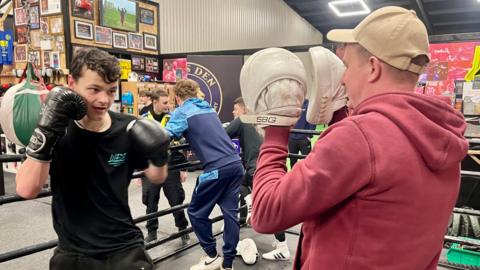 Two men stand inside a boxing ring in a gym. One has a black t-shirt, black gloves, black short hair, black tracksuit bottoms and a silver chain. He is holding his gloves up in preparation to strike. The other man faces him and his hands are covered by white cushioned pads. He is holding his hands in front of his face. This man is wearing a white cap, red hoodie and has a shaved head. In the background, two young men in tracksuits are talking to a police officer. There are lots of photographs framed on the wall of boxers.