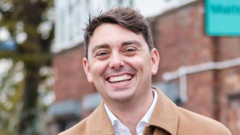 Adam Root is wearing a beige jacket over a white shirt. He is standing in front of a brick building and smiling. He has short dark hair.