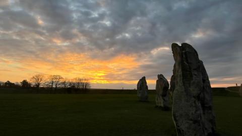 Some of the Avebury stones at dawn as the sun rises. An orange sun merging with light grey clouds.