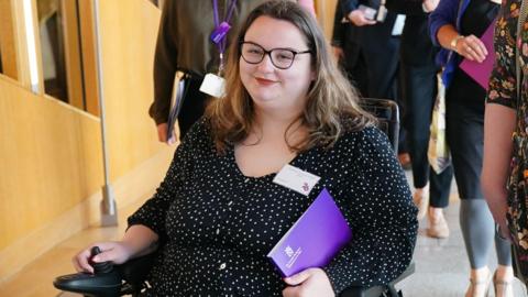 Kayleigh Kinross-O'Neill sits in a wheelchair. She has long brown hair and wears a black and white polka dot dress and carriers a purple book under her arm