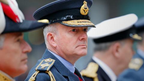 Chief of the Defence Staff Sir Richard Knighton (centre) looks wistfully away from camera while wearing formal military attire and a shiny black military hat while stood next to members of the Welsh Guards wearing bright red military uniforms.