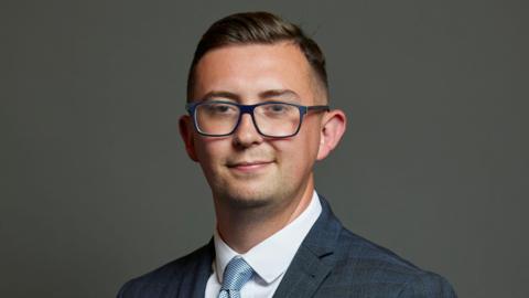 Connor Naismith, the Labour MP for Crewe and Nantwich, wears blue-rimmed glasses and has dark hair, parted on his left side. He has stubble on his chin. He wears a dark blue suit with slight light-blue checks on it with a white shirt and blue tie.