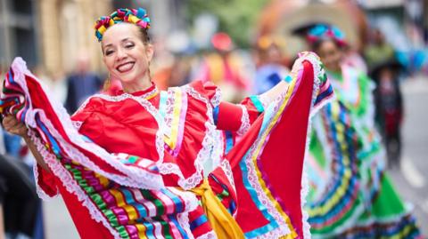 A woman with her brown hair pinned back holding up her traditional dress which is predominantly red with white lace and green, pink, yellow and blue ribbon during a parade. She wears large gold dangle earrings.