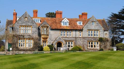A large historic looking boarding school with grey pointed bricks, terracotta tiled roof and lots of bay windows. There is a bright green manicured lawn in the foreground, and various climbing plants on the side of the building. 
