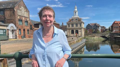 Alison Gifford stands on the King's Lynn Quayside with water behind her. On the quay is a small square stone building which is the Custom House. Alison is wearing a blue shirt and a string of white pearls. She has short grey hair and is smiling at the camera.