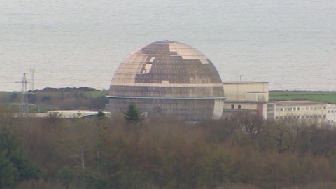 One of the Sellafield buildings, which is shaped like a sphere and covered in various shades of grey concrete. In the distance, the sea can be seen.