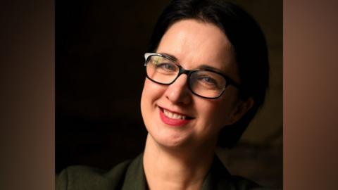Em Burnip has short black hair and is wearing a dark grey jacket. She has black framed colours and is wearing pink lipstick.