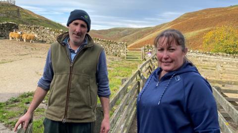 Daniel Wood is standing next to Sam Wood. Sam is a woman in her forties wearing a blue top with dark hair tied back. Daniel is wearing a jumper and a green jacket and blue woolly hat. Behind them is a stone wall with sheep up against it and in the distance are rolling hills on a sunny day.