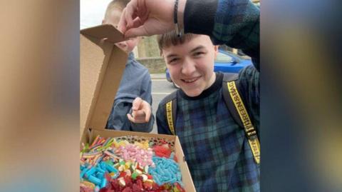 William Barron Hynes wearing a checked top and smiling as he holds open a cardboard box of sweets 