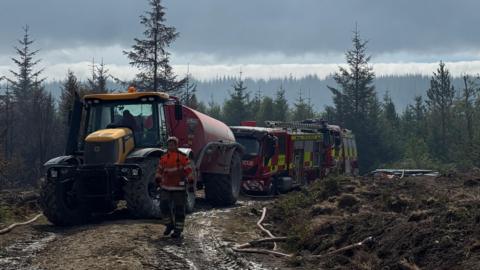 Man walking in front of a tractor and fire engines in moorland.