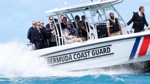 King Charles III sat on a Bermuda Coastguard boat, with a crowd of men stood around him