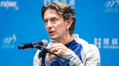 Thomas Frank sits in front of a blue background with a microphone before him