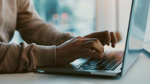 A person in a brown jumper typing on a laptop.
