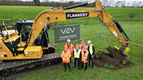 An aerial shot of six people wearing hi-vis jackets in a field, with a big yellow digger behind them.