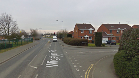 A Google Street View image of a main road's junction with a residential road, with houses on the right-hand side. 